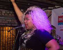 A woman with wild blonde hair with her right arm raised and her left on her hip sings into a mic in a dark brick room.