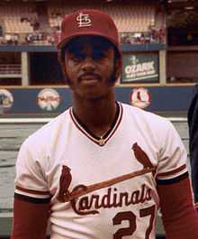 A dark-skinned man with long sideburns and a mustache wearing a white baseball uniform with "Cardinals" in red script across the chest and a red baseball cap