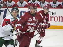 A group of three ice hockey players skate to the right of the image while looking to the left. The image is focused on the middle player, who is wearing the same uniform of the skater to his right, who is partially concealed by his teammate. Their opponent is to the left, with his team bench visible in the background