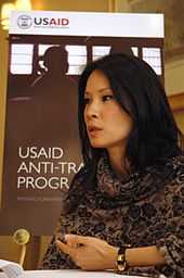 A photograph of a woman with long, black hair wearing a black-and-gold bracelet and facing the left, all in front of a sign reading "USAID"