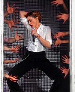 Madonna wearing a white shirt and black pant sings to a microphone, while to her sides several human hands can be seen.