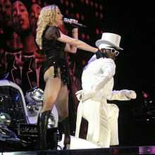 A faraway image of a female blond woman on stage in front of a white Speedster. The woman is wearing a white hat, black top and dancing. Two huge backdrops display two African American males in front of blinking lights. The one in the top backdrop looks towards the camera and smiles and the one in the bottom backdrop is looking down while wearing a white-rimmed sunglass.