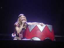 Image a blonde woman wearing red-and-white Circus outfits and using a black tiara. She is sat-down on the floor and is holding a michophone to her mouth, with her eyes closed.