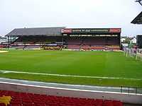 In the foreground, a grass football field. In the background, a two-part seating area. Both parts are covered and are connected, but the roofs are distinctly different.