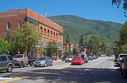 A wide undivided street with cars traveling in both directions going past a large brick building. In the rear is a forested mountain