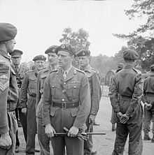 General officer with gloves and cane in hand, talking to a sergeant, surrounded by other officers and men