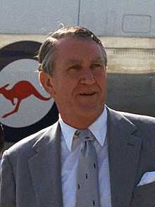 Photograph of a man aged about fifty, he has a weathered face and greying hair parted on the right. He wears a suit and tie; behind him can be seen part of a large aircraft with a kangaroo logo.