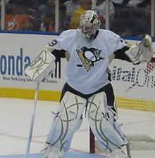 Hockey player in white uniform and goaltender's gear. He stands in front of the goal, legs shoulder length apart, and has raised his right arm as if to catch a puck, while his left arm holds a hockey stick.