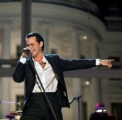 A man standing, holding a microphone with stand with his right hand and with his left hand extended, wearing a two-piece black suit, with a white shirt, a necklace and silver watch.