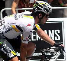 A road racing cyclist wearing a black and white jersey with yellow and green trim. His bicycle is only partly visible.