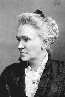 Chest high portrait of a middle aged woman wearing a dark dress and white shirt, hair up in a bun
