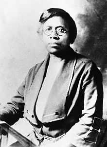 An African American female with short dark hair, round eye glasses, and a solemn expression sitting down and facing the camera.