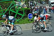 A pair of road racing cyclists in the team's black, white, and blue jersey, riding around a traffic island that features a large green sculpture of a bicycle. Spectators and motor vehicles are visible behind roadside barricades in the background.