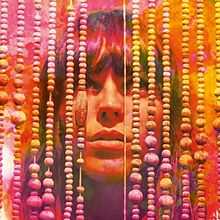 A woman standing stares upwards behind a series of hanging beads; the centre of her face is visible. The image is overlaid with alternating shades of red, pink, purple, orange and yellow.