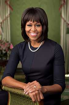 Michelle Obama facing forward, smiling, clad in black dress and single strand pearl necklace resting bare right forearm and both hands on a brocaded sofa armrest.