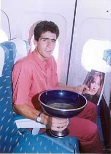 A man sitting in an a blue seat, holding a trophy and a magazine