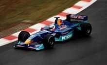 A figure, wearing a helmet with a white and aqua design, is driving a Formula One car that is of a dark blue and aqua colour scheme. He is holding both hands on a steering wheel, turning right.