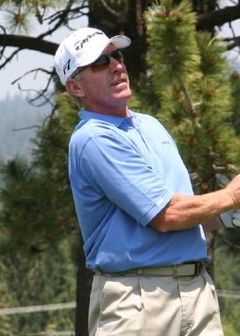 A man wearing a sky blue polo shirt, khaki pants, sunglasses, and a white cap stands with his right forearm across his stomach.