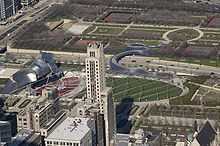 View from a height of a green park with a bandshell surrounded by curved shiny metal, with a trellis over a large green lawn. A curved metal bridge crosses a horizontal street to another park. In the foreground, skyscrapers block part of the view.