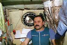 A man, dressed in blue work clothing, seen in a small cubicle. On the walls around him can be seen a sleeping bag, children's drawings, technical manuals and stained insulation. A small porthole in the centre of the wall behind him shows the nose of the Space Shuttle Atlantis and the blackness of space.