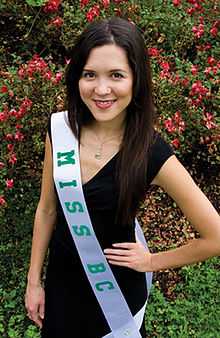 A photograph of a woman looking at the viewer and smiling while wearing a black dress, an necklace, and a white sash with green letters on it