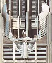  A stylized figure of a male human with outstretched arms and head tilted slightly forward, wearing a winged and crested helmet, mounted on the facade of a building