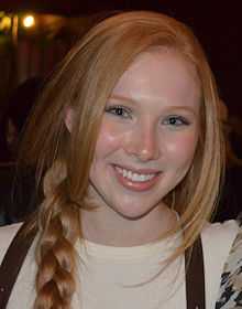 An eighteen-year-old redheaded female is wearing a beige shirt with her ponytail draped over he right shoulder; she is smiling into the camera.