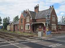 View across the tracks and level crossing of a fine 19th century station building in red brick and with stone mullioned windows, under a roof of Welsh slate.  The platform has gone.