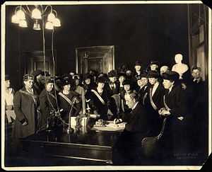 A man sits at a desk sigining a document while a large group of women watches