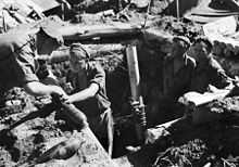 A black and white image of soldiers standing in a fire pit around an indirect fire support weapon