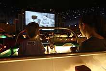 A rear view photograph of a woman and a boy sitting in a booth, the boy watching a film on a cinema screen in the distance and the woman looking down