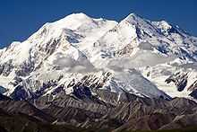Mount McKinley, covered in snow at the top, and green and brown near the bottom. Some wispy clouds are in the middle.