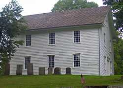 A two-story white building with a pointed roof, seen from the side, with gravestones in front, similarly austere