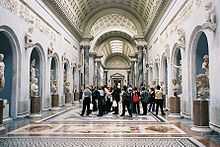 Photo of a gallery in the museum. It is in the Classical style and has a wide arched roof with skylights. The colour scheme is blue-grey and white with a polychrome marble floor. The walls of each side of the gallery have a row of large niches in which stand marble statues. Between the niches are plinths supporting smaller portrait sculptures.