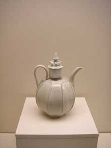 A white teapot with an almost perfectly spherical body and a large, cylindrical cap in the center which is topped with a small crown shaped embellishment. Several vertical lines are glazed into the body of the pot.