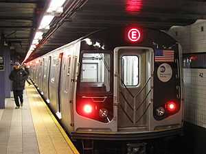 The end of a subway train with a red E on the end sits next to a platform with a person walking away.