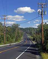 A road, with wooden telephone poles and lines on either side, descending from the camera into a wooded area and then ascending in the background