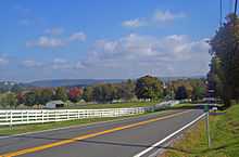 A rolling landscape with hills in the distance, a line of trees some of which are turning red, and fields in the foreground. A white fence separates them from a road that stats at bottom left and then passes between trees at the right center before cresting. At the bottom right is a small green sign with white numbers on a metal stand.