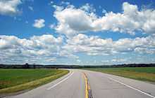 A paved two-lane road curving to the left into the distance through a large grassy area below a blue sky filled with cumulus clouds