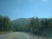 A two-lane highway in a forested area. To the right of the highway is a sign indicating that a hill is ahead. In the distance is a large mountain completely covered with trees.
