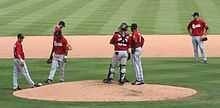 Seven baseball players wearing black caps, red jerseys with "Sounds" written across the chest, and gray pants standing around the pitcher's mound during a game