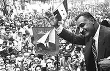 A man wearing a suit and tie with his upper body jutting out, waving his hand to crowds of people, many dressed in traditional clothing and holding posters of the man or three-striped, two-star flags