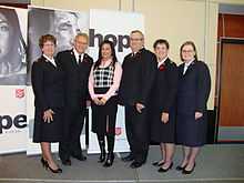 A photograph of six standing, smiling people, five of whom are wearing black uniforms with the sixth wearing a pink dress shirt
