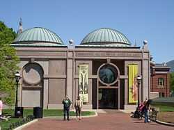 A one story symmetrical granite building with two green domed roofs either side of center. The entrance door is on the right and a similar design is on the left made out of a slightly darker granite.