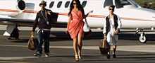 From left to right: a young black man, a young brunette in a red dress and a young white man walking on a jet landing space and carrying luggage. All are wearing sunglasses and in their background is an already landed jet.