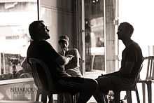 A black-and-white photograph of three men sitting on chairs facing each other and wearing shorts and t-shirts all next to another empty chair