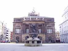  A Neoclassical building seen end-on.  It has two storeys with a central colonnaded portico and the dome can be seen above the roof.  In the foreground is an elaborate monument