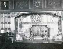 A black and white photograph of a fireplace with ornate mantel set in wood-paneled wall with similarly ornate carvings above it