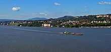 A city on a slope over a body of water with larger buildings at the left and center right. There are low hills in the distance, and a large ship crossing the body of water at the bottom of the image