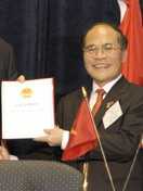 a smiling nearly bald man, wearing glasses, a suit and a red tie while holding up a piece of paper